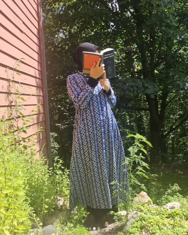 Person in a long blue dress holding a book in front of their face outdoors.