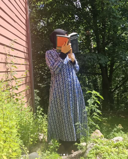 Person in a long blue dress holding a book in front of their face outdoors.